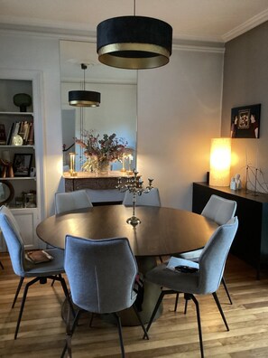 Ein Essbereich mit einem runden Tisch, grau gepolsterten Stühlen, einer schwarzen und goldenen Pendelleuchte sowie einer Wandlampe.