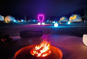 A fire pit with flames, a heart-shaped neon light, and dome-shaped structures in the background.