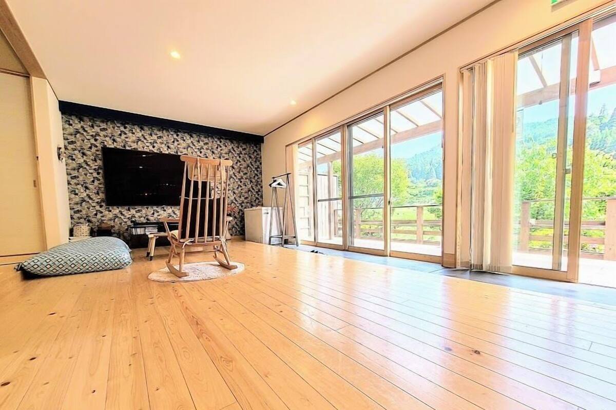 A spacious room with a wooden floor, a large window with a view of greenery, a mounted TV, and a rocking chair.