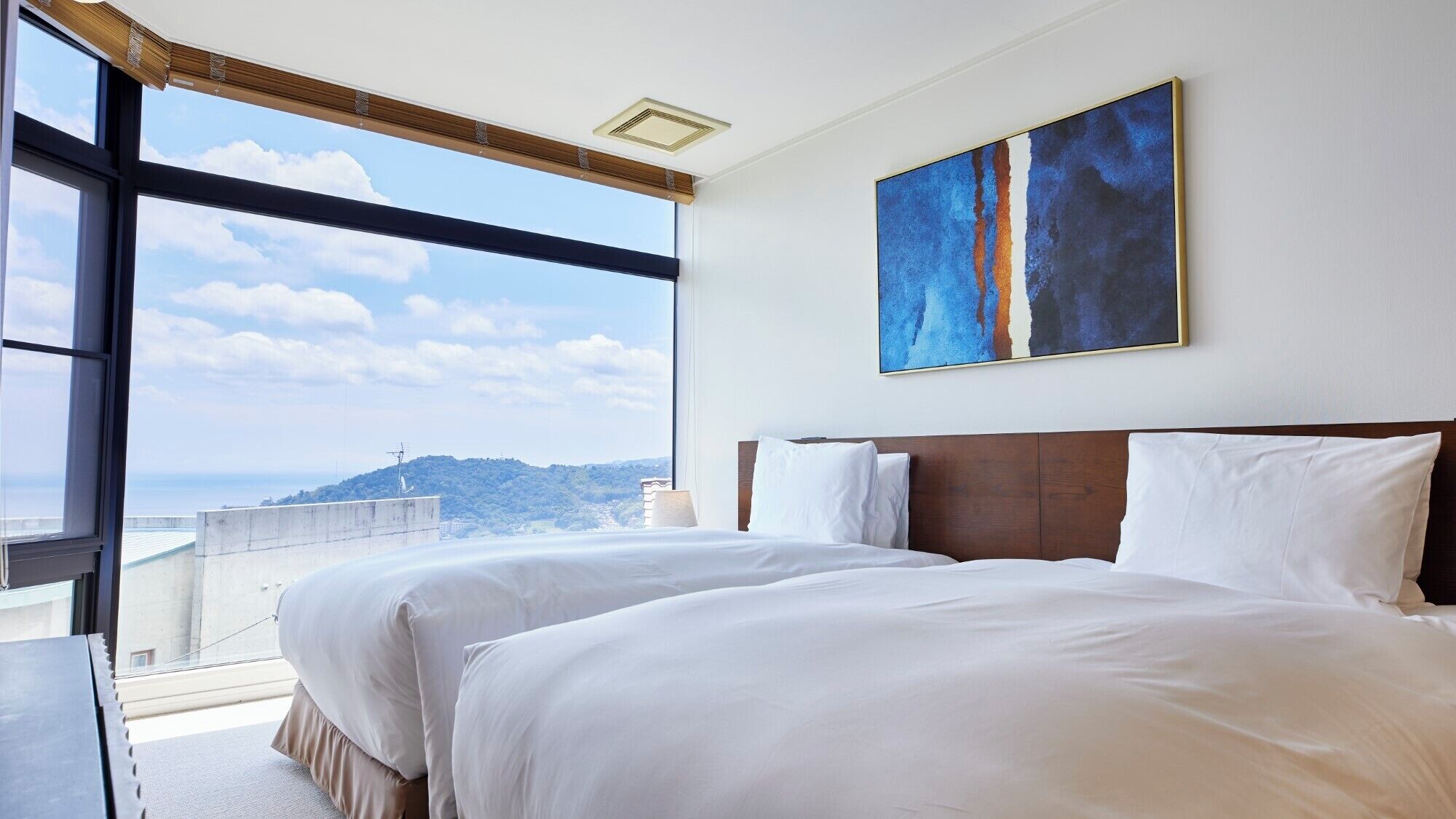 A hotel room with two beds, a large window, and a painting on the wall.