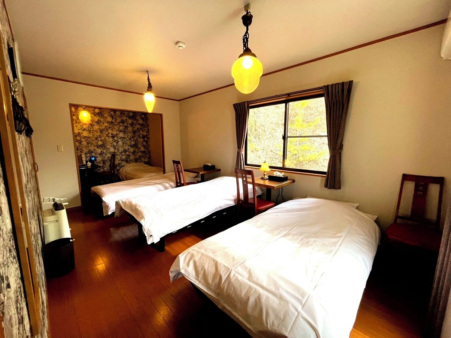 A hotel room with two beds, a small table, a chair, a window with curtains, and a ceiling light fixture.