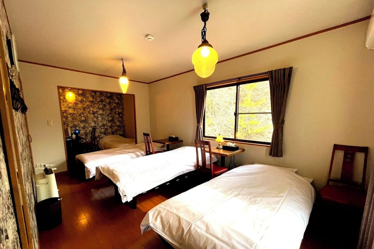 A hotel room with two beds, a small table, a chair, a window with curtains, and a ceiling light fixture.