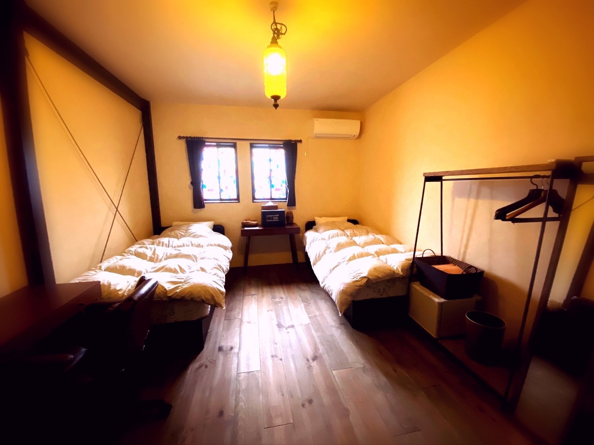 A room with two beds, a desk, a chair, a hanging rack, and a ceiling light.