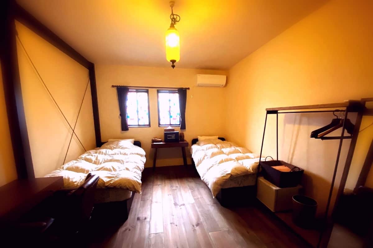 A room with two beds, a desk, a chair, a hanging rack, and a ceiling light.