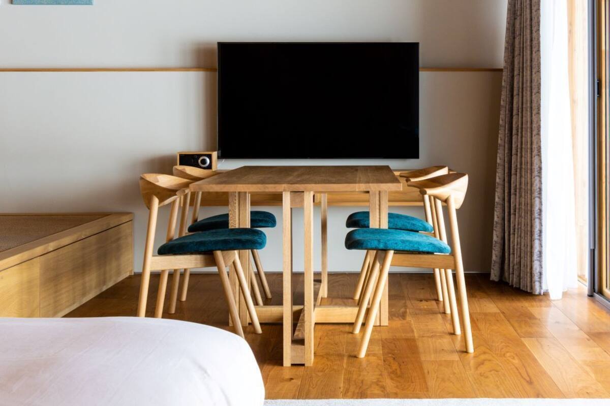 Una habitación con techo de madera, un televisor de pantalla plana en la pared, una mesa de comedor de madera con sillas, una cama con ropa blanca y una ventana con cortinas.