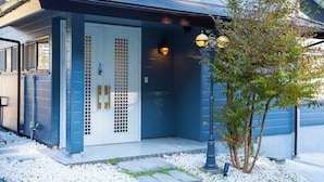 A building with a blue exterior, a white door with a grid pattern, and a small tree in front. - Maigre Fields (Atami)