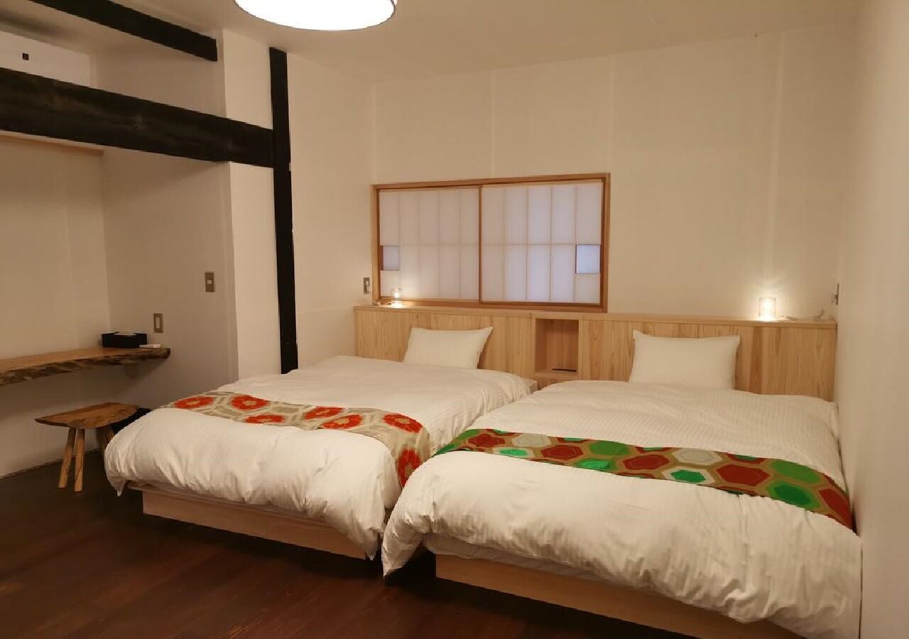 Una habitación con dos camas, un cabecero de madera, una mesita pequeña y una ventana con persianas.