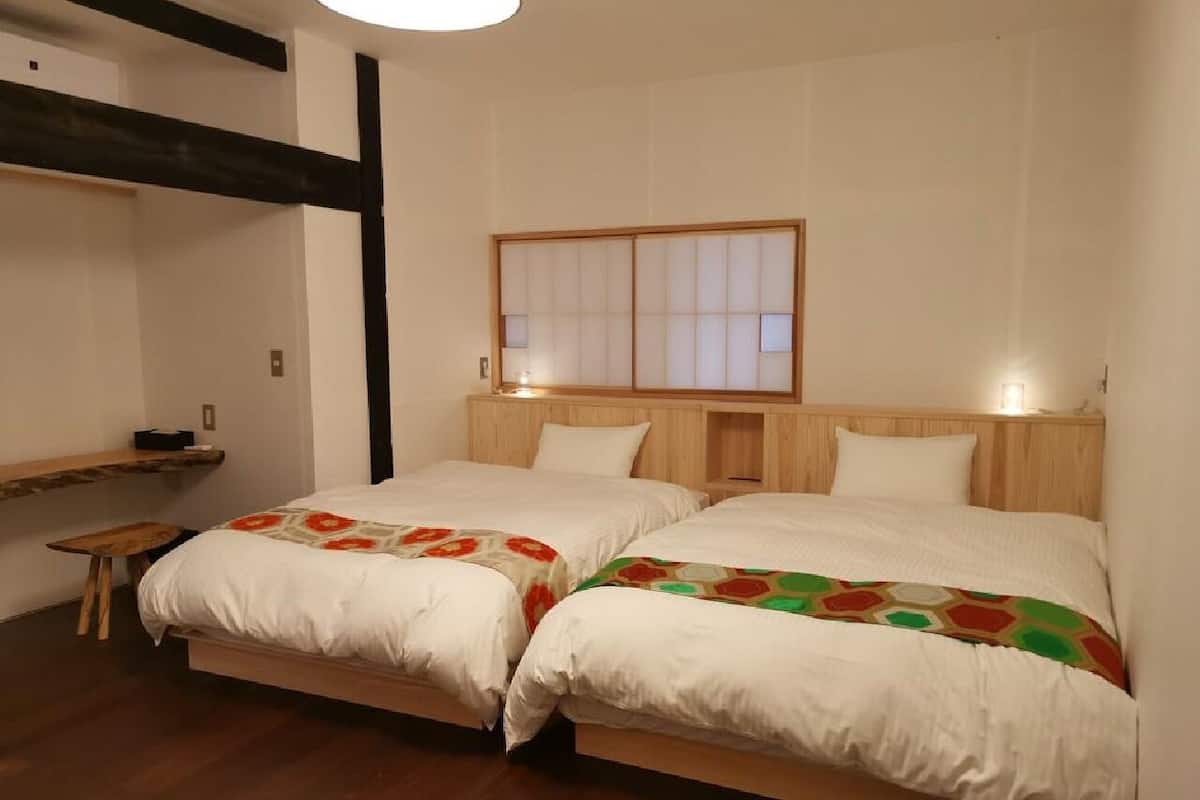 Una habitación con dos camas, un cabecero de madera, una mesita pequeña y una ventana con persianas.
