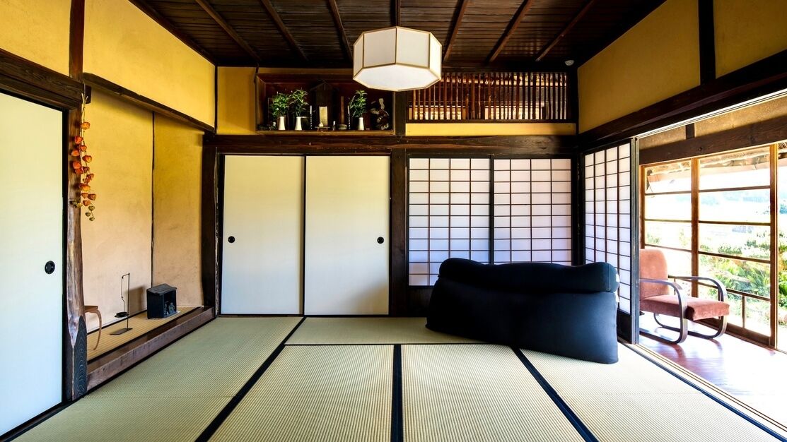 Una habitación japonesa tradicional con suelo de tatami, un sofá negro, una lámpara colgante y vistas a través de un shoji.