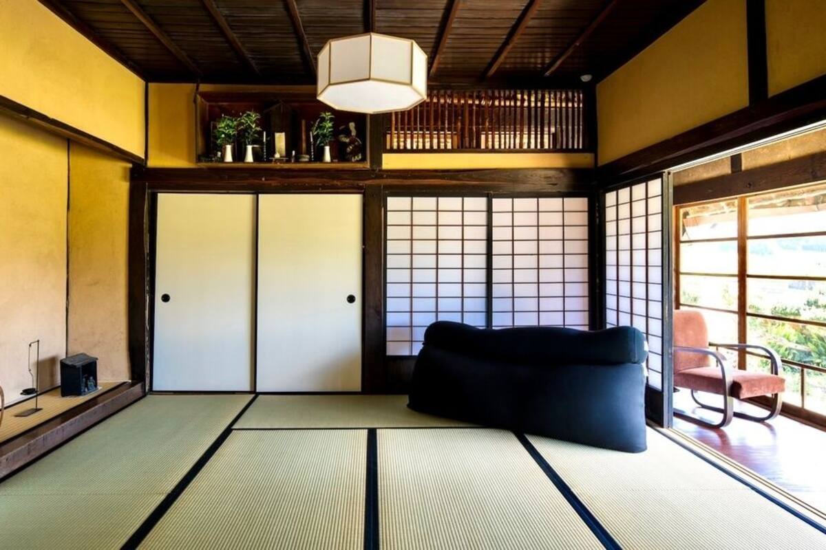 Una habitación japonesa tradicional con suelo de tatami, un sofá negro, una lámpara colgante y vistas a través de un shoji.