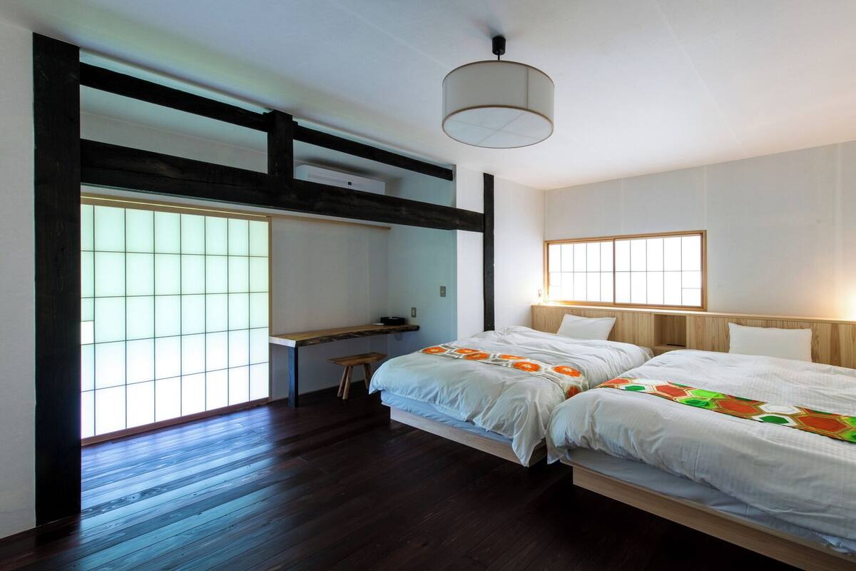 Una habitación con dos camas, un escritorio, una silla, un ventanal y una lámpara colgante.