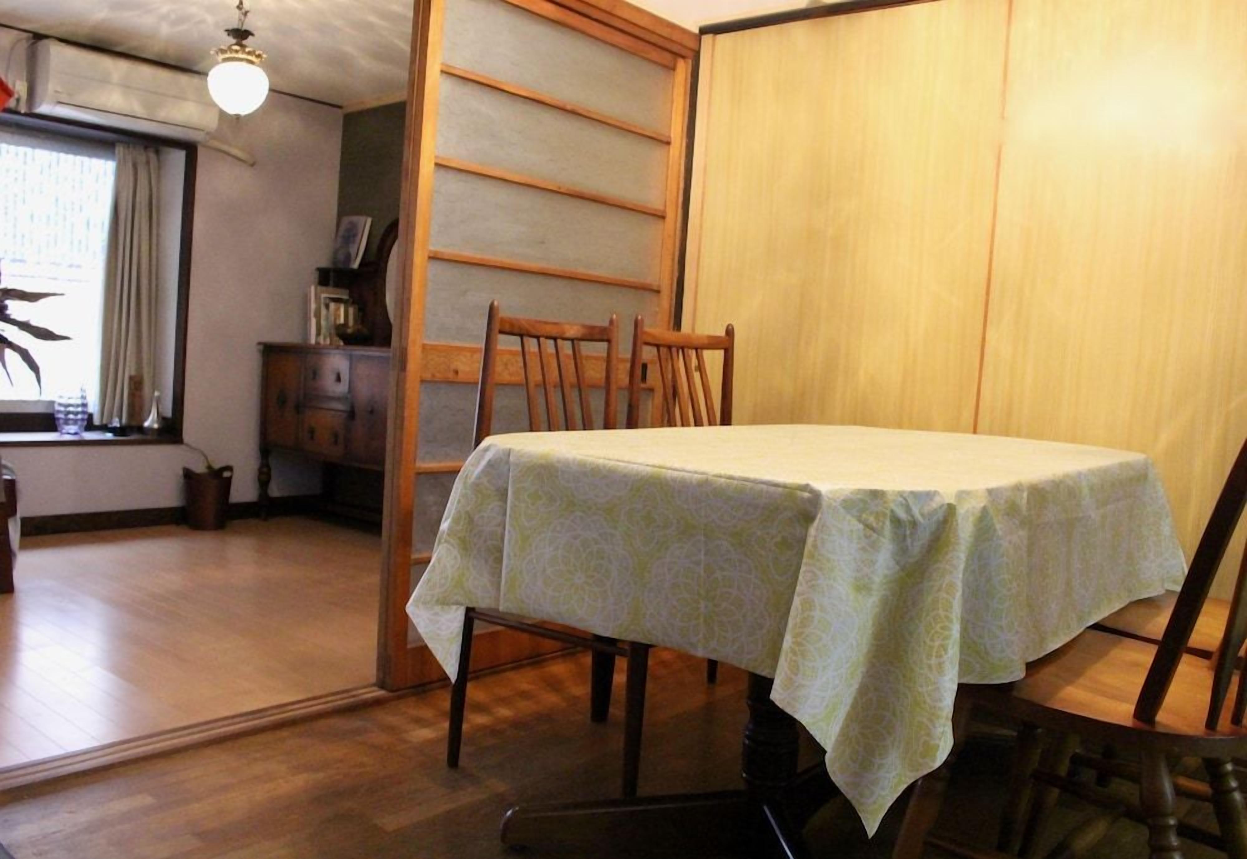A dining area with a table covered by a patterned tablecloth, wooden chairs, and a sliding door in the background.