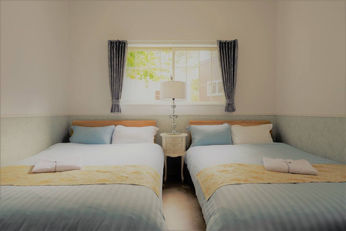 A double room with two beds, a bedside table with a lamp, and a window with curtains.