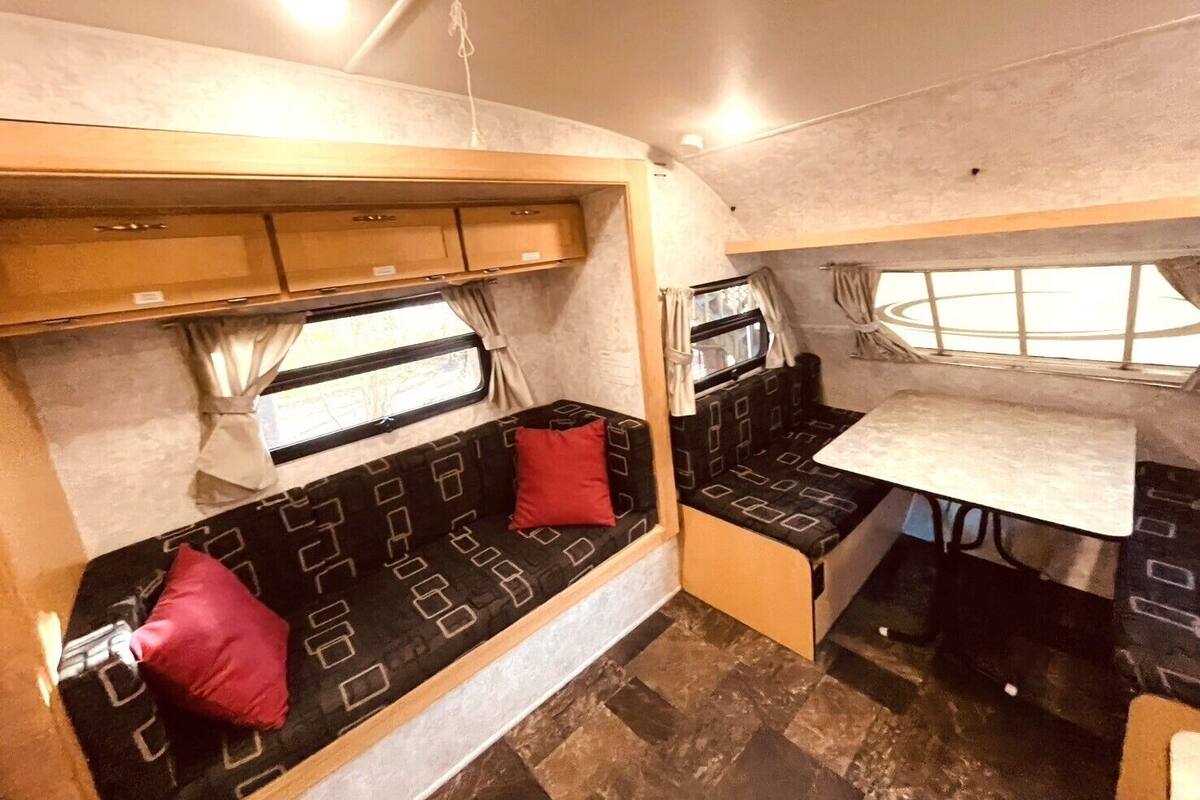 A compact RV interior with a kitchenette, a seating area with cushions, and a window providing natural light.