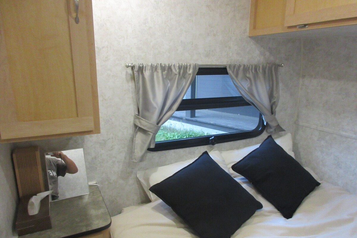 Una habitación pequeña y funcional con cama, ventana con cortinas, lavamanos y espejo.