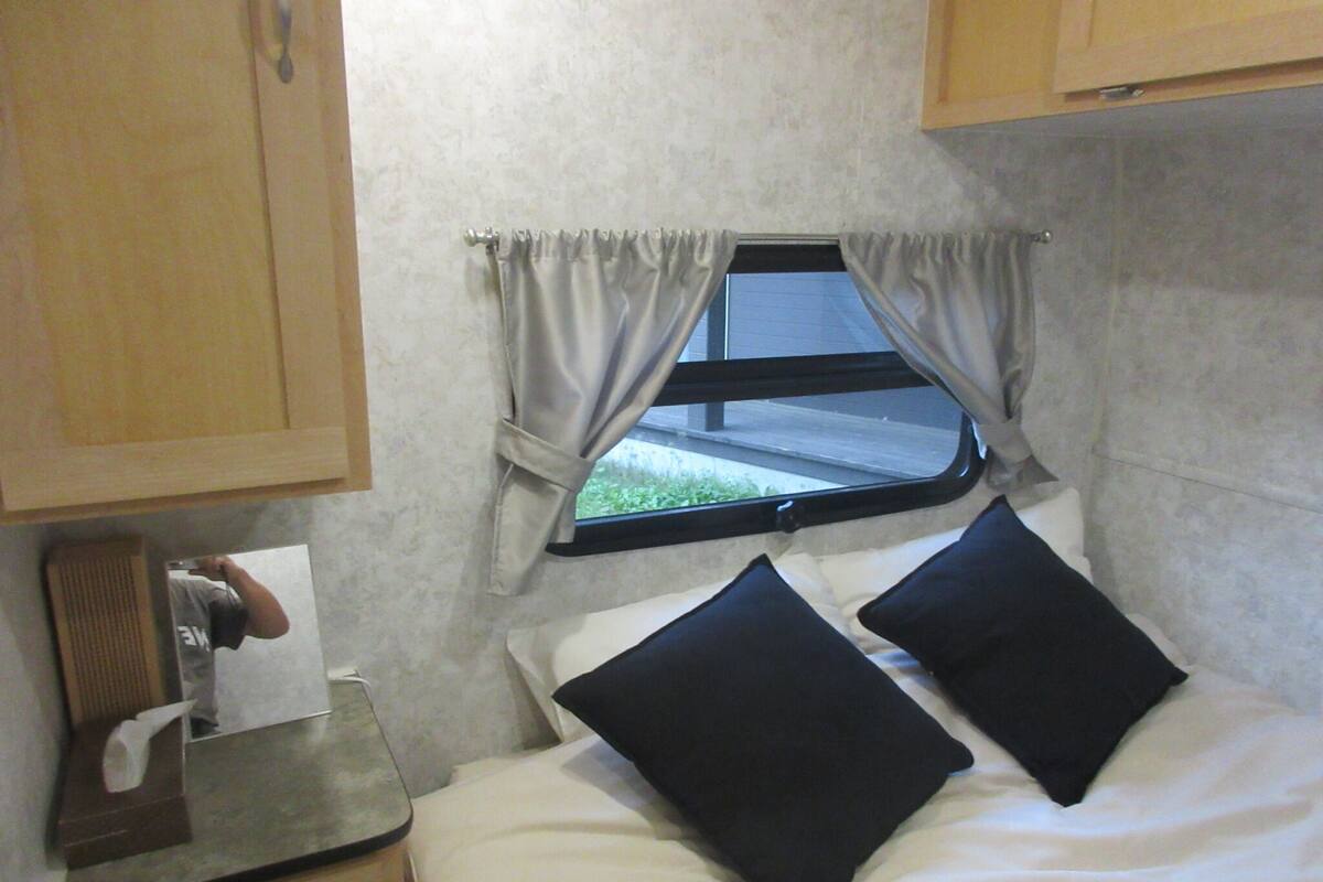A small, compact room with a bed, a window with curtains, a sink, and a mirror.