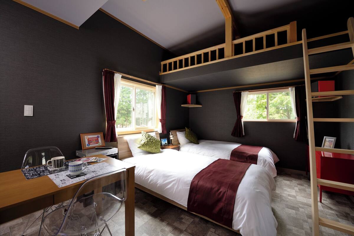 A room with two beds, a desk, a chair, a ladder, and a window.