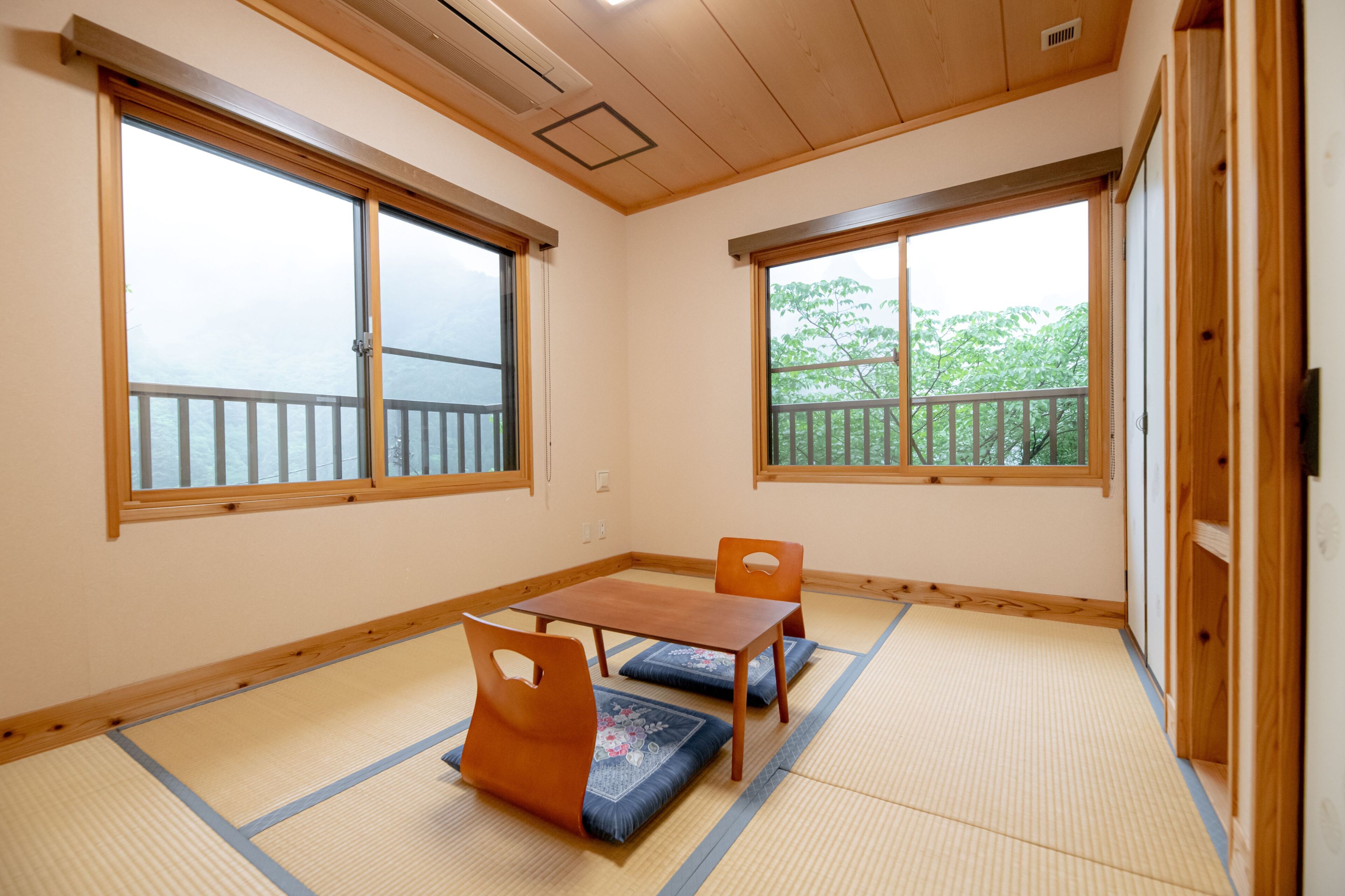 A room with tatami flooring, a wooden table, two chairs, and large windows with a view of greenery.