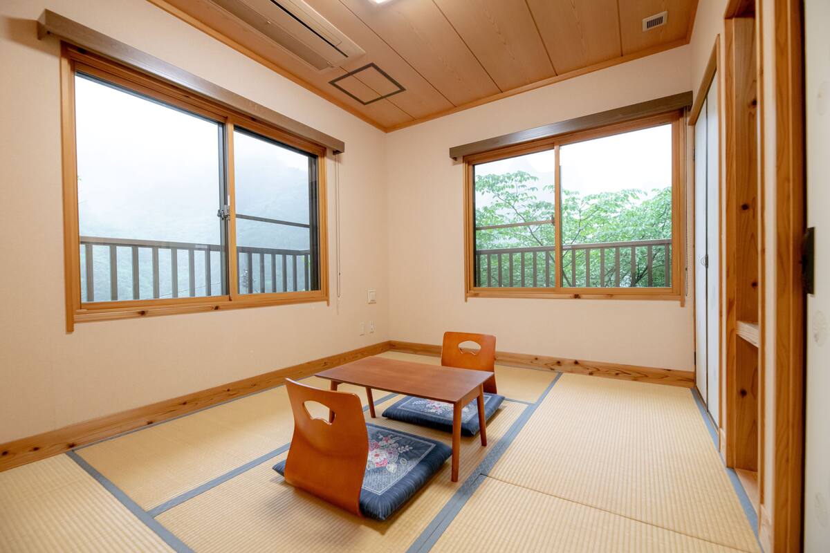 A room with tatami flooring, a wooden table, two chairs, and large windows with a view of greenery.