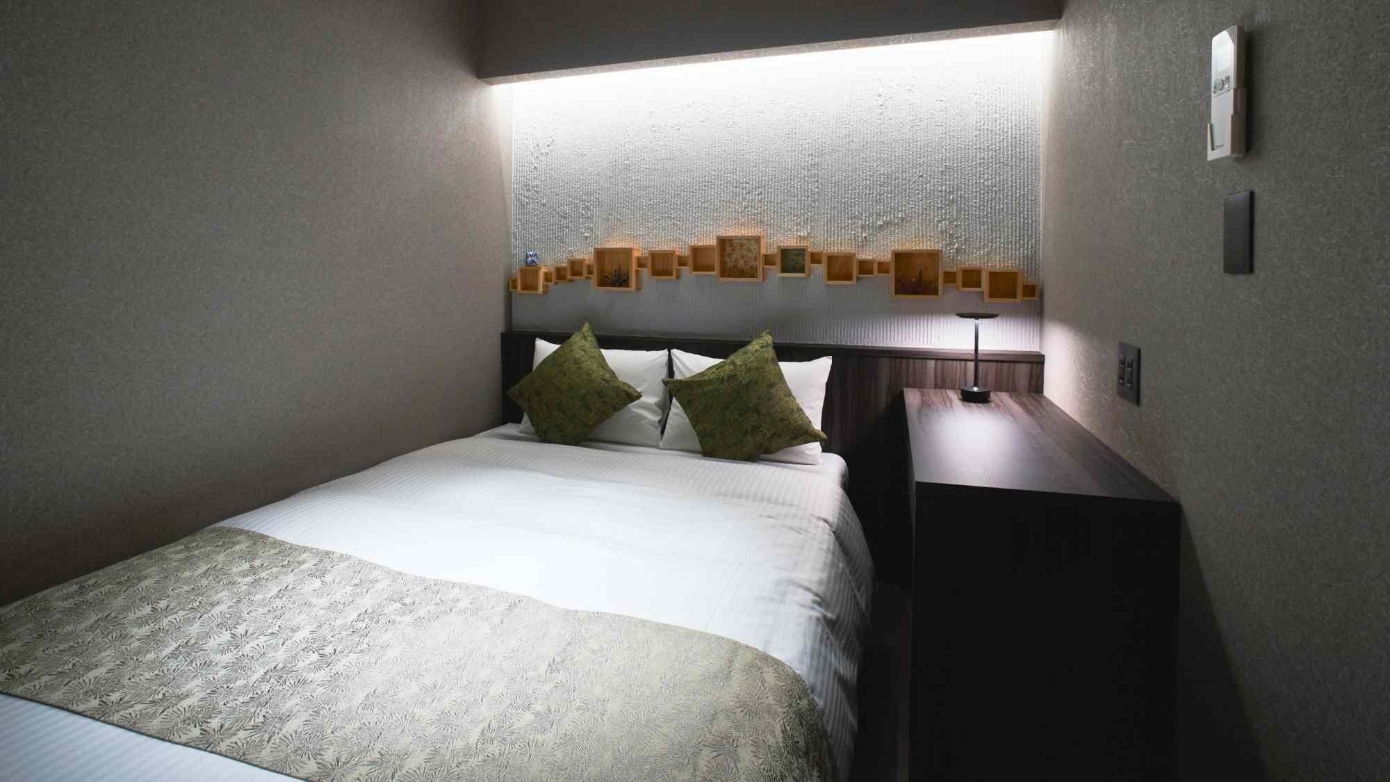 A hotel room with a bed, two pillows, a bedside table with a lamp, and a wall-mounted light fixture.