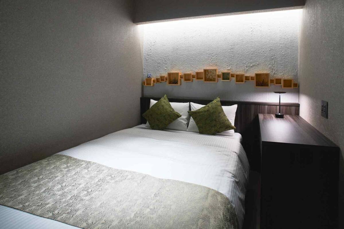 A hotel room with a bed, two pillows, a bedside table with a lamp, and a wall-mounted light fixture.