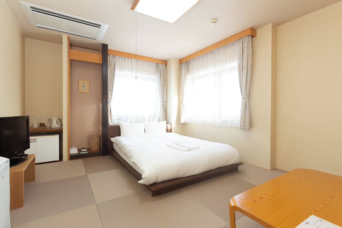 A hotel room with a bed, a desk with a television, a small refrigerator, and a window with curtains.