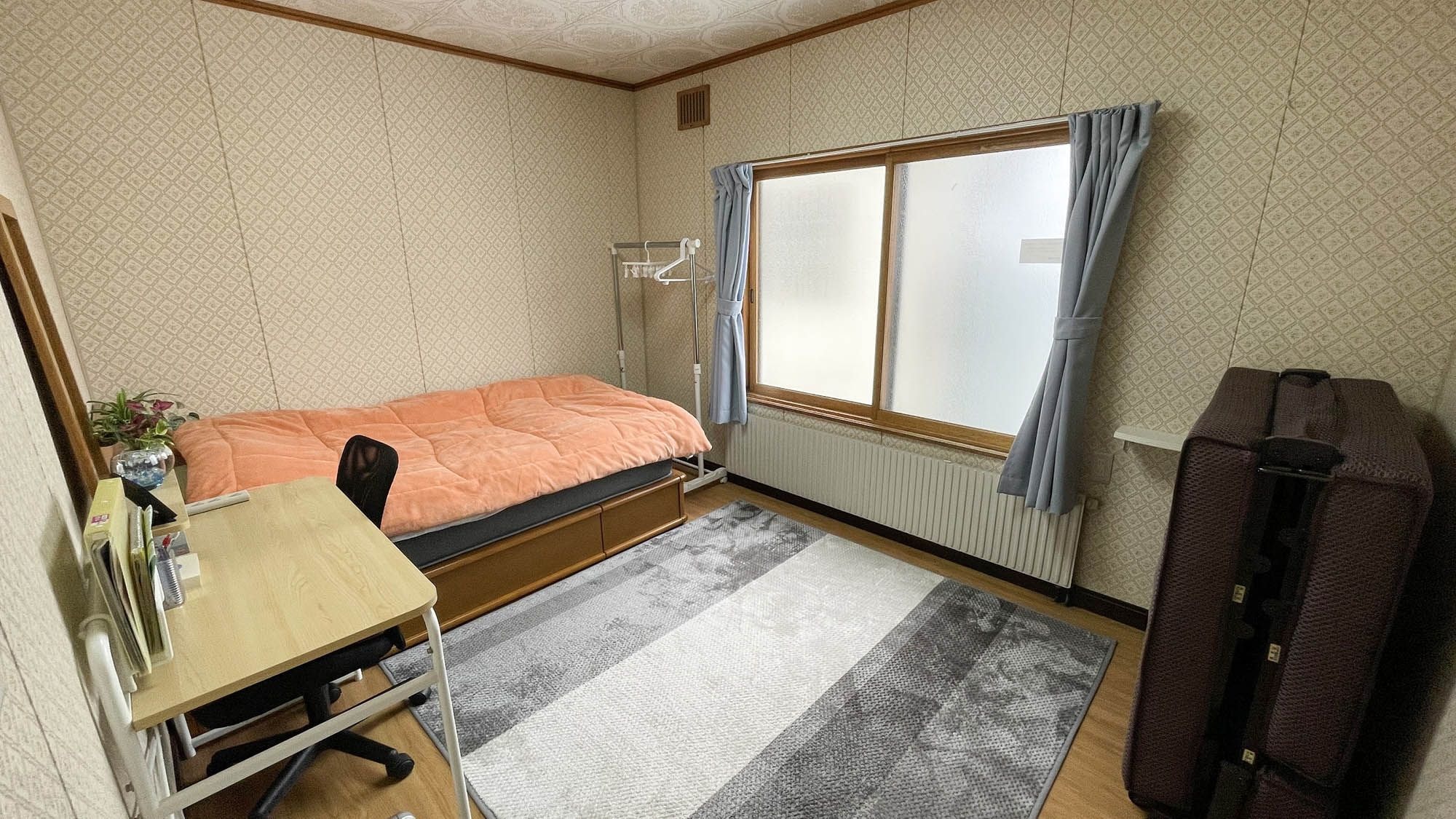 Habitación pequeña con una cama individual, un escritorio, un radiador, una ventana con cortinas y una maleta.