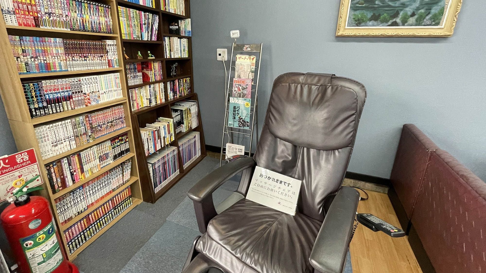 Una habitación con un sillón de cuero, una estantería llena de libros, un extintor y un cuadro en la pared.
