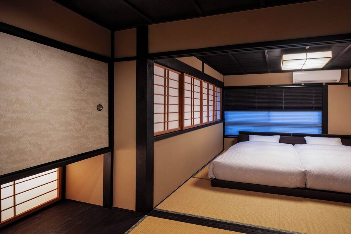 A traditional Japanese-style bedroom with tatami flooring, a bed with white bedding, sliding shoji doors, and a view of the outside through a window with blinds.