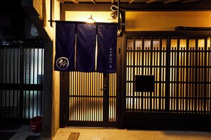 A traditional Japanese gate with a sliding door, a hanging blue cloth with a logo, and a red bucket on the ground. - Rinn Gojo Mibugawa East (Kyoto)