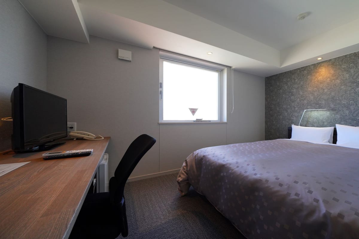 A hotel room with a bed, a desk with a television and phone, a window, and a patterned wall.