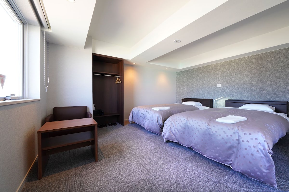 A hotel room with two beds, a desk, a wardrobe, and a window.