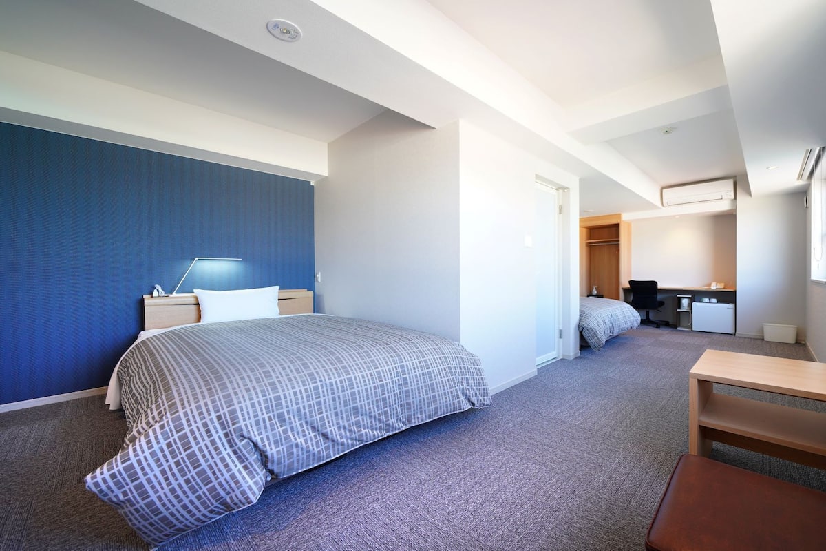 A hotel room with a bed, bedside table, lamp, desk, chair, and a small table.