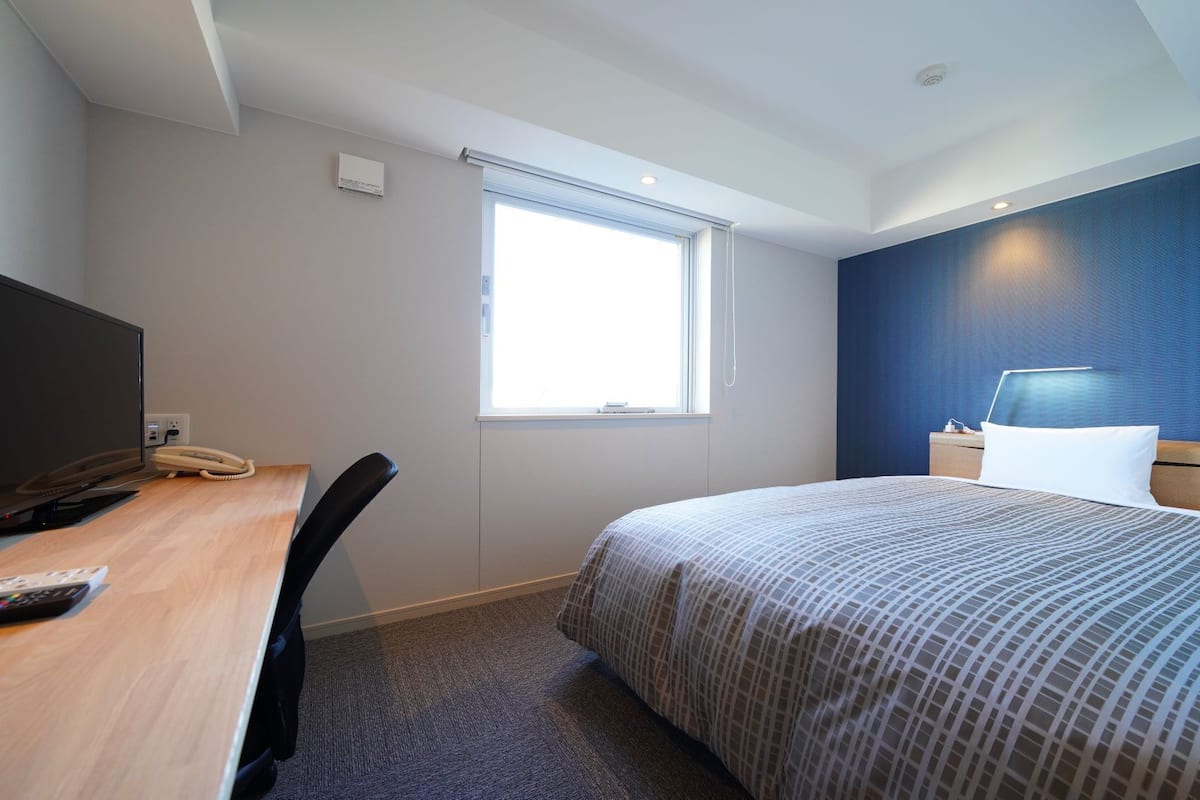 A hotel room with a bed, a desk with a TV, a chair, a window, and a nightstand with a lamp.