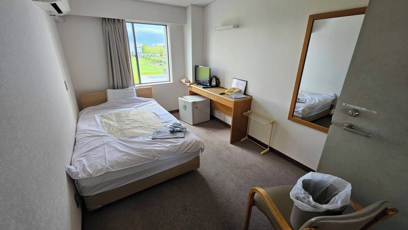 A hotel room with a bed, a desk, a computer, a mirror, and a view of the outdoors.
