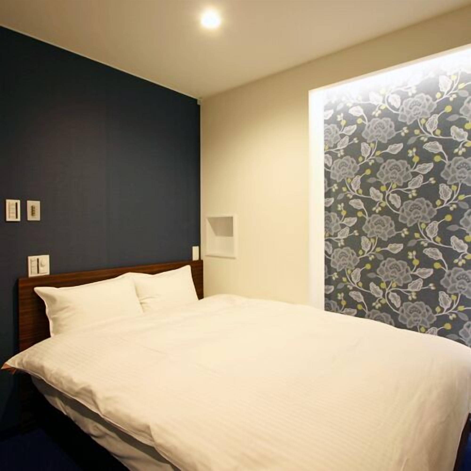 A neatly made bed in a hotel room with a dark wall and a floral wallpaper on one side.