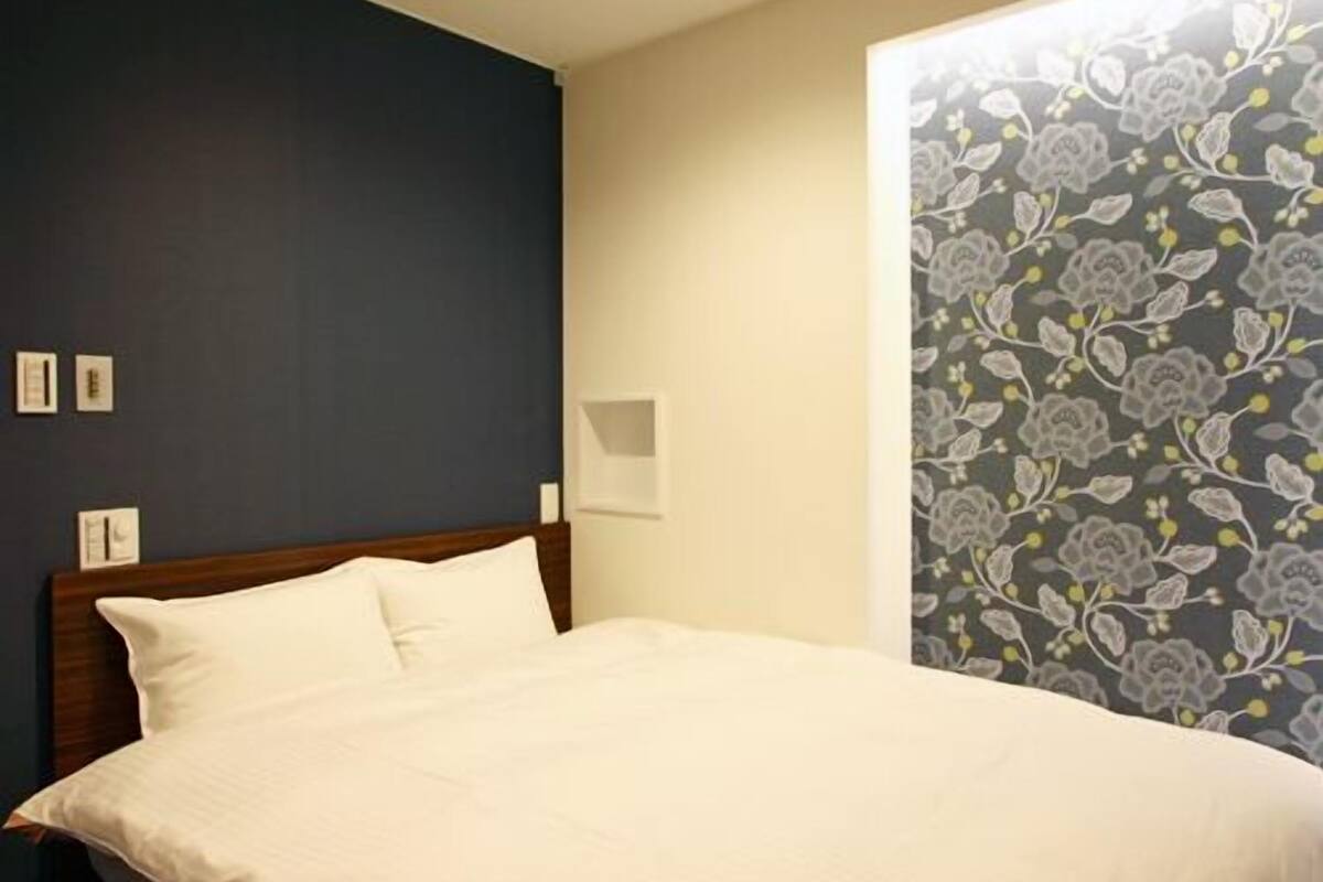 A neatly made bed in a hotel room with a dark wall and a floral wallpaper on one side.