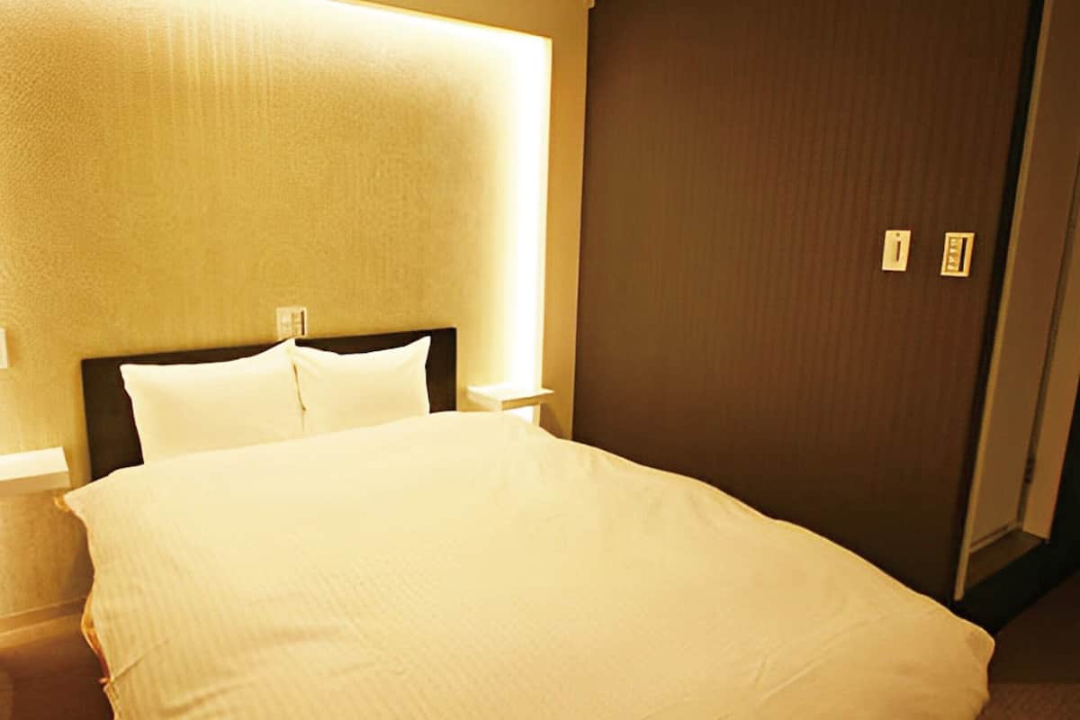 Hotellihuone, jossa on suuri sänky, yöpöytä, seinään kiinnitetty valaisin ja lukittava ovi.