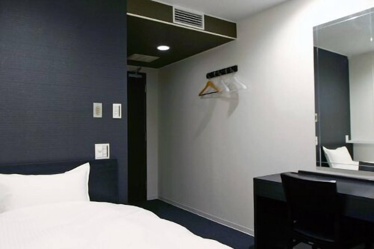 A hotel room with a bed, a desk, a chair, a mirror, and a coat rack with hangers.