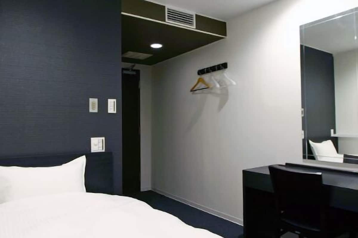 A hotel room with a bed, a desk, a chair, a mirror, and a coat rack with hangers.