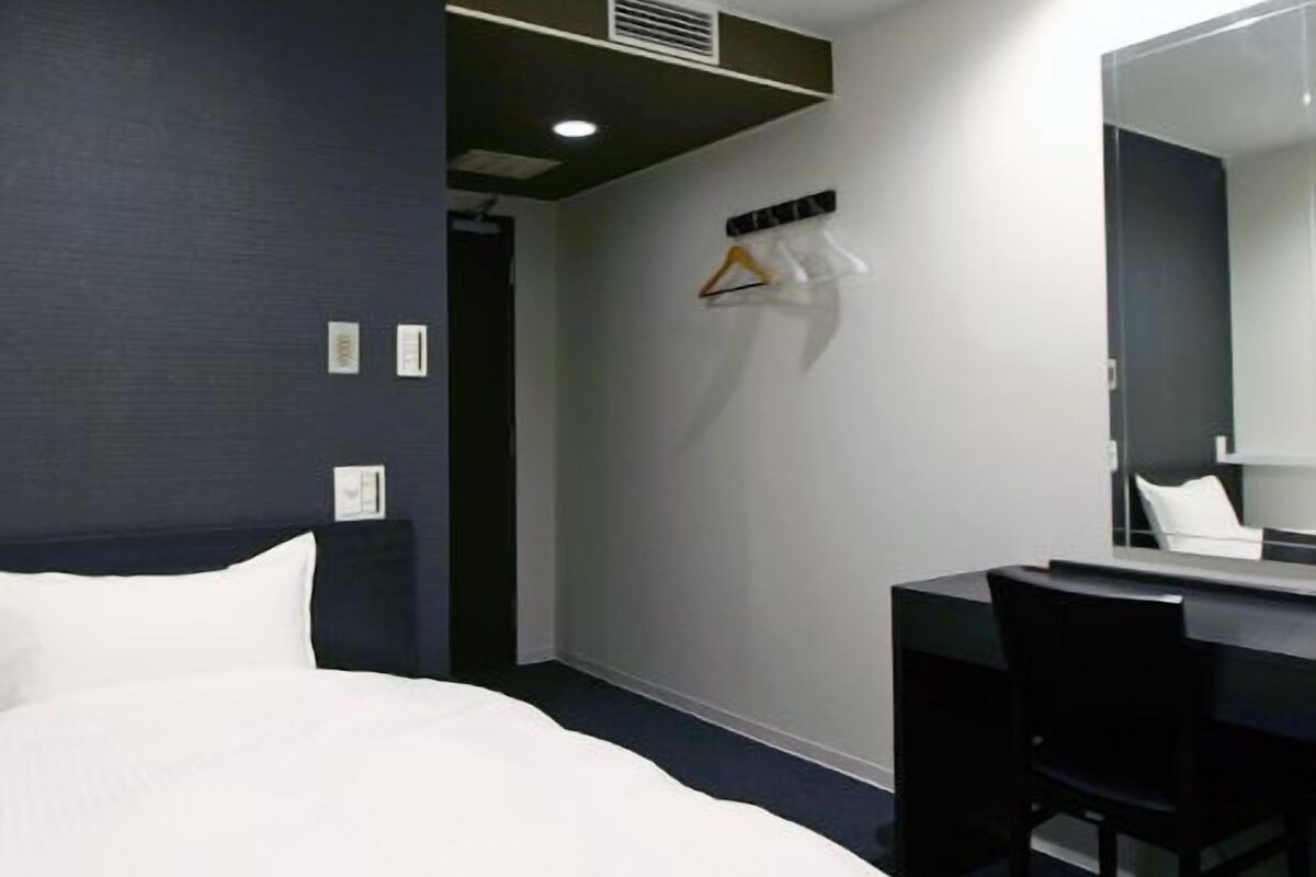 A hotel room with a bed, a desk, a chair, a mirror, and a coat rack with hangers.