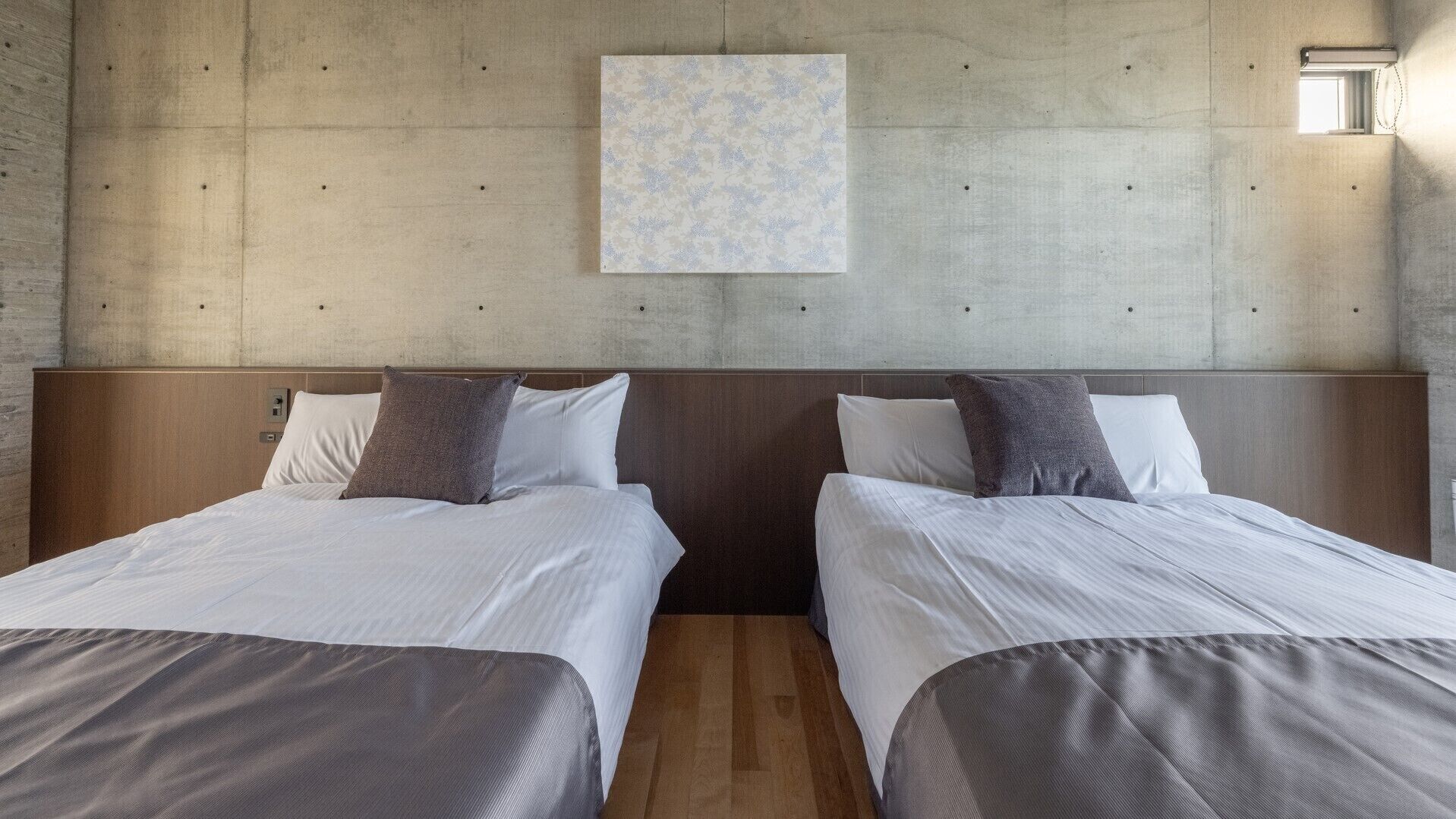 Habitació d'hotel amb dos llits, una paret de formigó i una finestra petita.