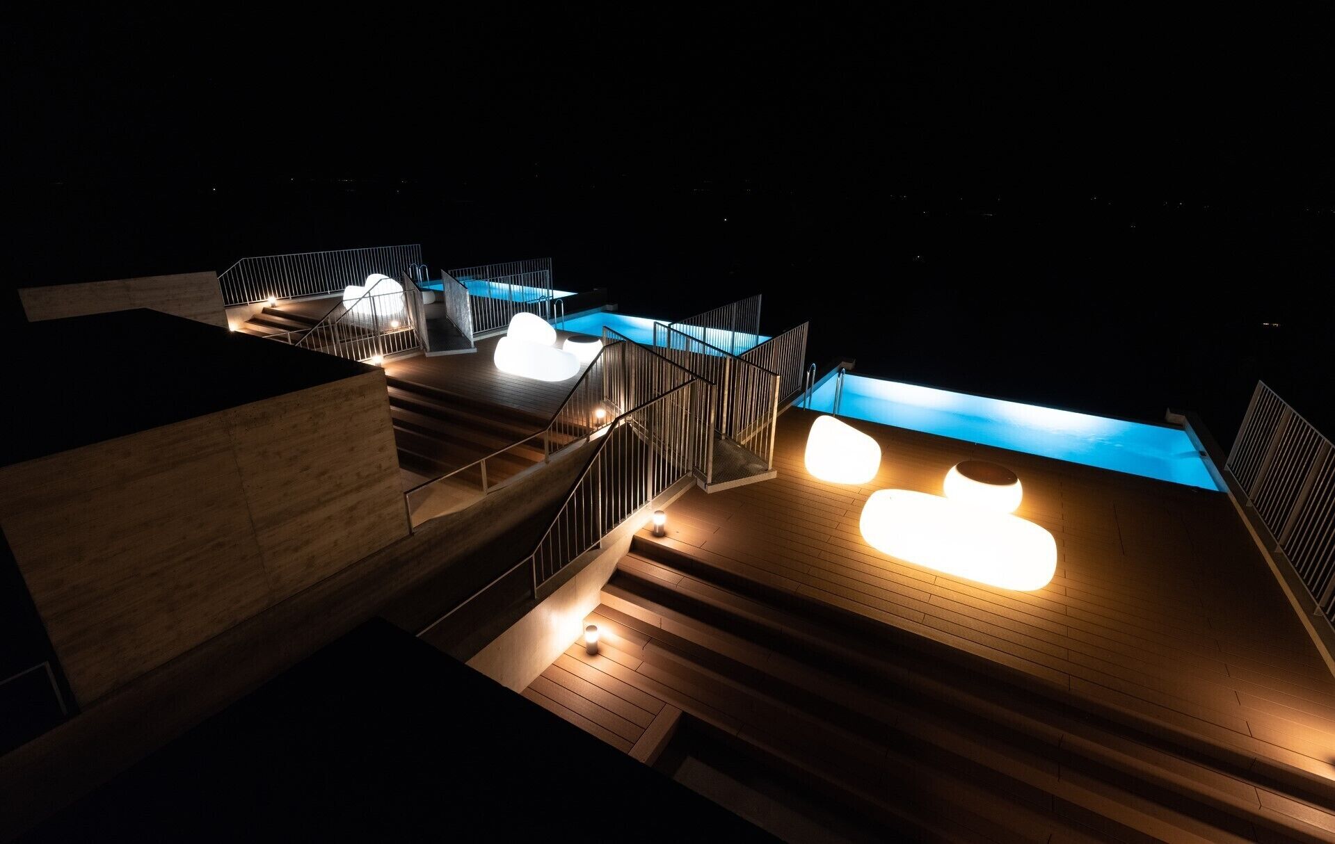 A modern outdoor pool area with a wooden deck, metal railings, and illuminated pool lights.