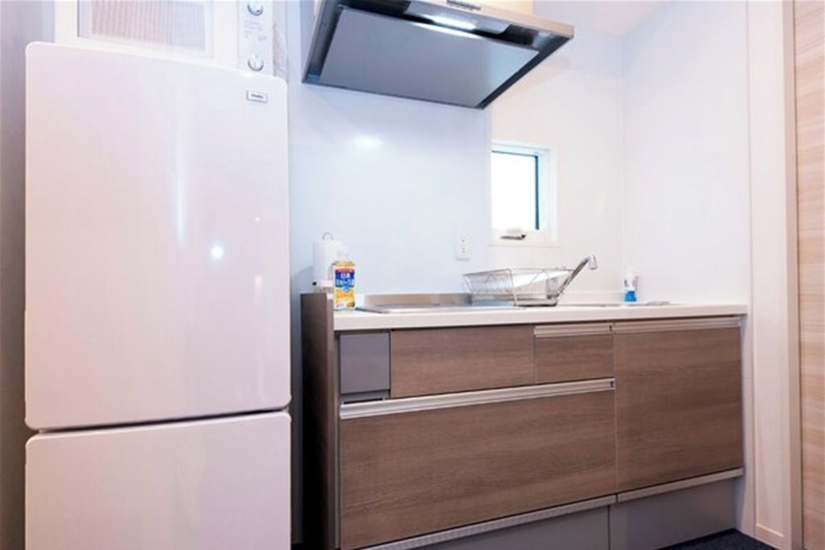 A compact kitchen with a white refrigerator, a microwave, and a built-in oven. There is a window above the sink, and a countertop with a sink and a trash bin.