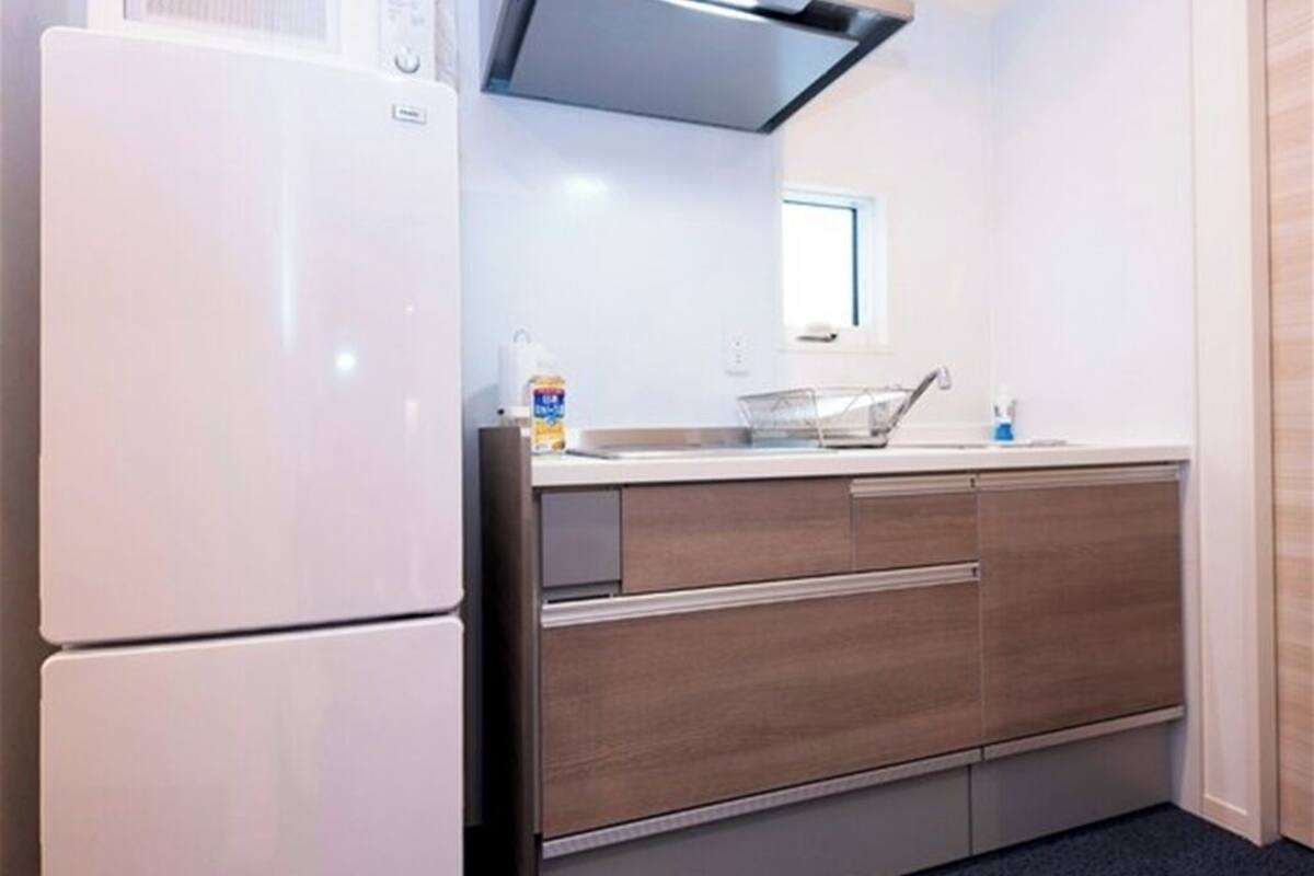 A compact kitchen with a white refrigerator, a microwave, and a built-in oven. There is a window above the sink, and a countertop with a sink and a trash bin.