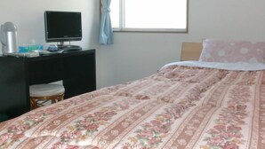 Ein Schlafzimmer mit einem Bett, einem Schreibtisch mit Computer, einem Stuhl und einem Fenster mit Vorhängen.