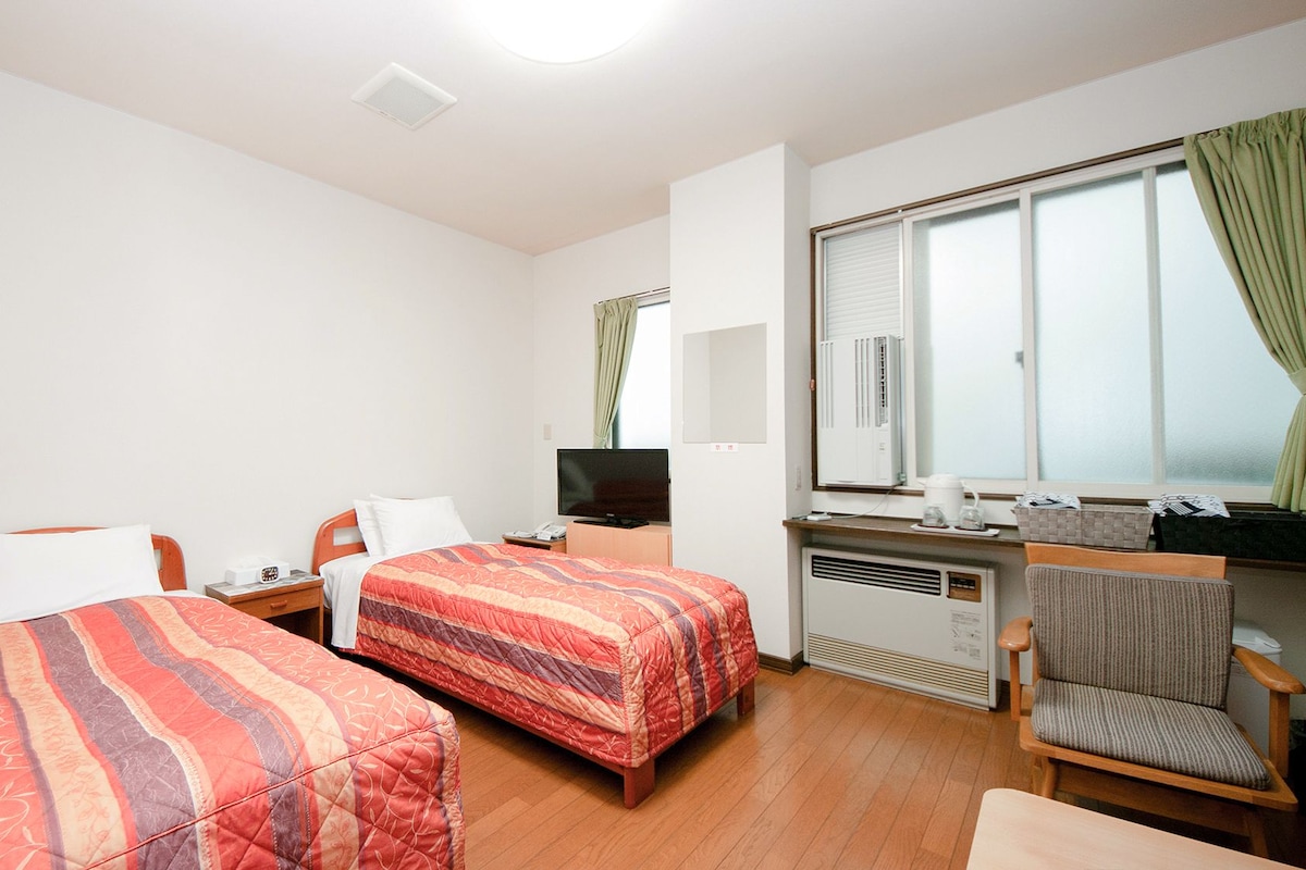 A hotel room with two beds, a TV, a desk with a computer, a chair, and a window with curtains.