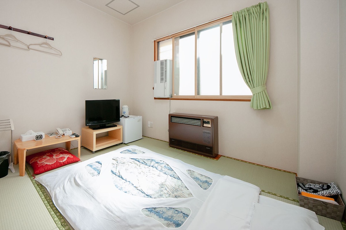 A hotel room with a bed, a TV, a heater, a window with curtains, and a wall-mounted heater.