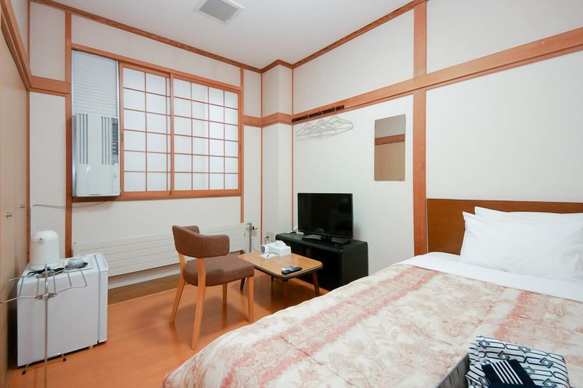 Una habitación de hotel con una cama, un escritorio con silla, un televisor y una ventana con persianas.