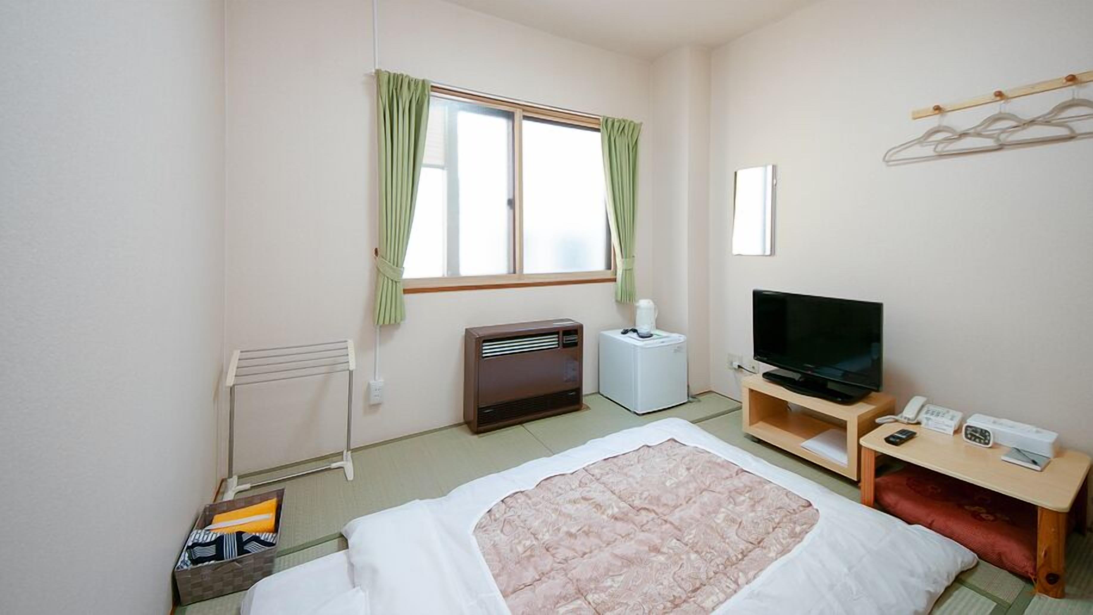 A hotel room with a bed, a TV, a heater, a radiator, a window with curtains, and a small table with a phone and a remote control.